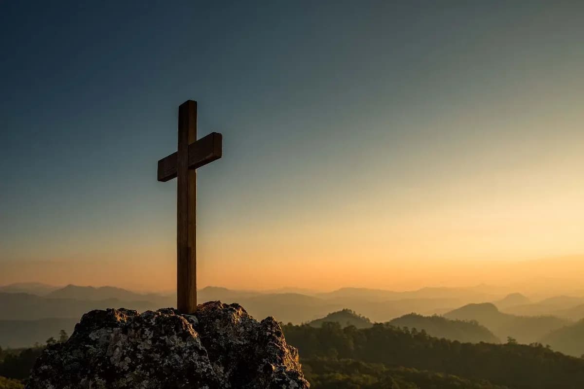 Wooden cross overlooking a mountain landscape at sunset.